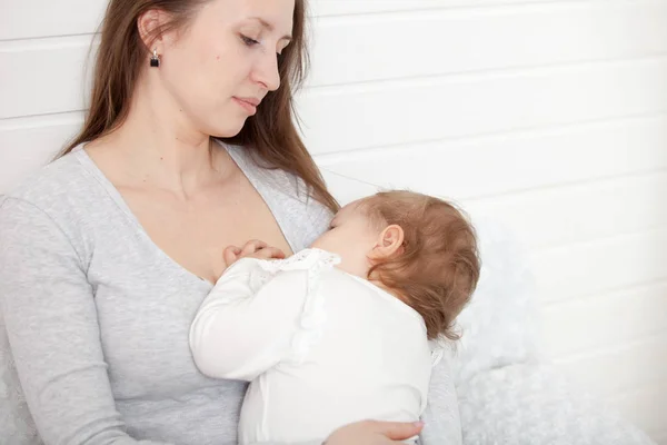 A young woman feeds the baby's chest, sitting on the bed — Stock Photo, Image