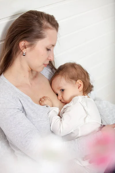 A young woman feeds the baby's chest, sitting on the bed — Stock Photo, Image