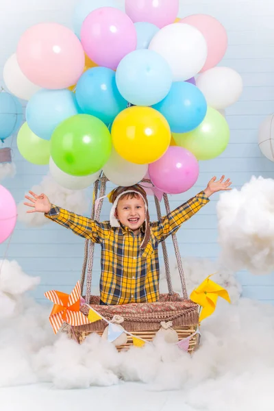 Una niña pequeña está sentada en una canasta de globos de aire caliente en el parque fingiendo viajar y volar con un sombrero de piloto para un concepto de creatividad o imaginación . — Foto de Stock