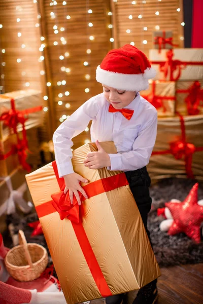 A joyful boy in a light shirt and Santa Claus hat takes a gift o — Stock Photo, Image
