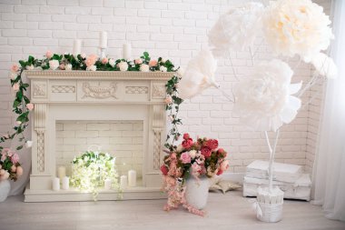 Zarif bir şömine, beyaz çikolata rengi, çiçekler ve mumlarla süslenmiş. Şömine kapısı olan romantik bir bahar süslemesi, büyük kağıt çiçekler ve yapay çiçekler..