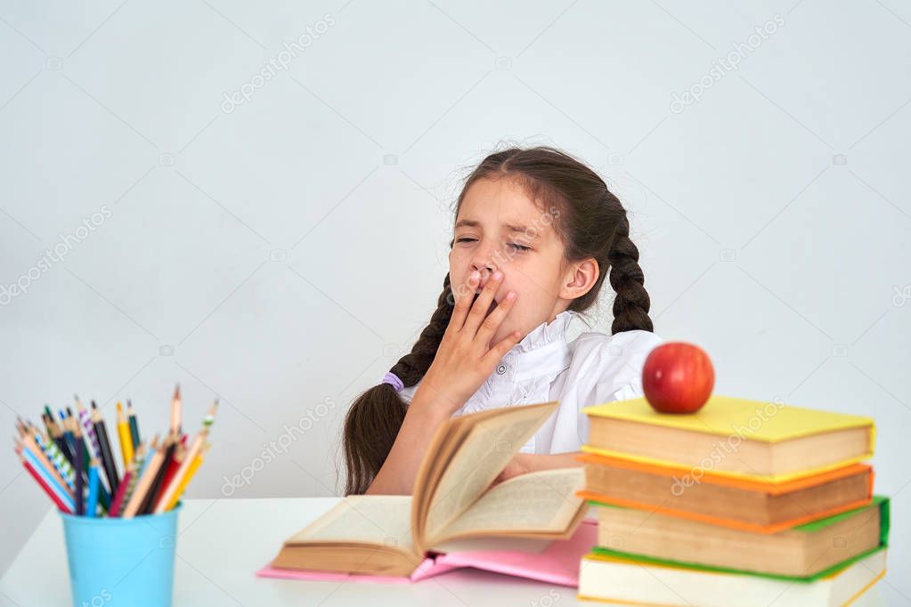 Overworked, exhausted by homework, a child, a school girl, with two pigtails, sits at a Desk and yawns. Tired of school and homework. On the table are textbooks, stationery, and an Apple.