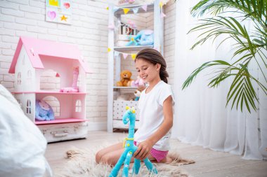 Açık pijama giymiş, kumaştan bir oyuncakla, zürafayla çocuklarının odasında oynayan mutlu şirin bir kız. Oda ışık ve oyuncaklarla dolu. Sevimli kız mutlu ve dikkatsizce gülümsüyor ve oynuyor.