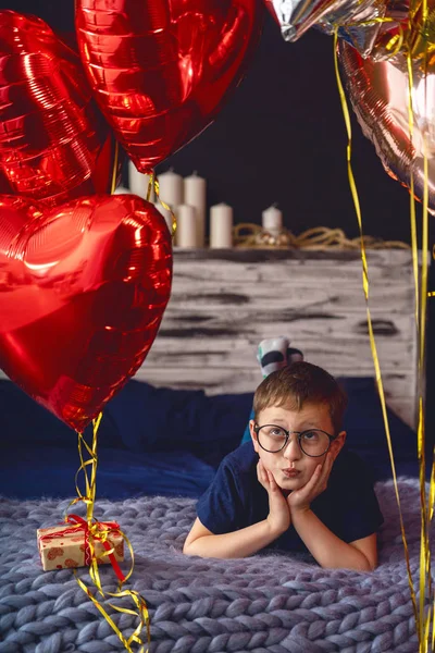 一个心事重重 戴着眼镜的男孩躺在床上 抬头看着一堆气球 一个7岁的孩子 梦幻般地看着球 等着度假呢等待的概念 — 图库照片