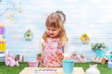 Mutlu Paskalyalar! Küçük tatlı bir kız fırçayla bir Paskalya yumurtası boyuyor. Çocuk mutlu bir şekilde yeşil çimenlerde oturuyor ve Paskalya tatiline hazırlanıyor. Tavşanlı Paskalya süslemesi..