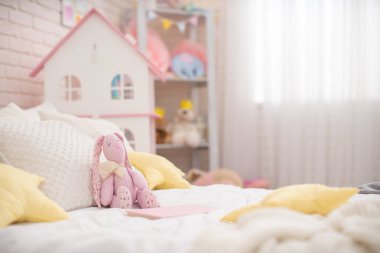 Kumaştan yapılmış el yapımı peluş tavşan çocukların odasındaki rahat bir yatağa oturur. Çocuk odası, Dollhouse ve bir sürü oyuncak var. Yatakta bir sürü yumuşak yastık yıldızları var. Boşluğu kopyala.
