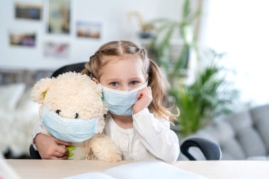 Maskeli küçük bir kız, oyuncak ayıya sarılıyor, üzgün görünüyor, e-öğrenme konsepti, karantina ve kendini izole etme sırasında. Covid-19 koronavirüs salgınından dolayı uzaktan öğrenim.