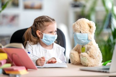 Maskeli küçük bir kız, oyuncak ayıyla ödev yapıyor, deftere yazıyor, dizüstü bilgisayar kullanıyor, e-öğrenme kavramları kullanıyor, karantina ve kendini tecrit sırasında. Covid-19 koronavirüs salgınından dolayı e-öğrenim.