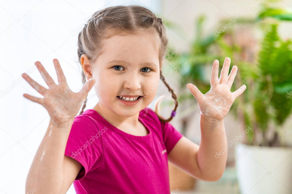 Cute little girl washes her hands in the bathroom . The child shows lathered palms in the frame. Protection against infections and viruses during epidemics and pandemics. Background blur.