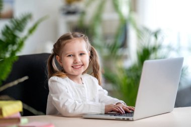 Küçük kız bilgisayarını evden öğrenmek için kullanıyor. Çocuk mutlu bir şekilde gülümser ve uzaktan bilgi alır. Dijital konsept e-öğrenme, salgın sırasında, karantina ve kendini izole etme. Uzak e- öğrenme.