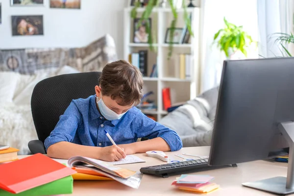 Boy in face mask with computer does his homework during a coronavirus quarantine. concept of online education. Social distance, self- isolation. Distance learning due to virus, flu, epidemic COVID-19