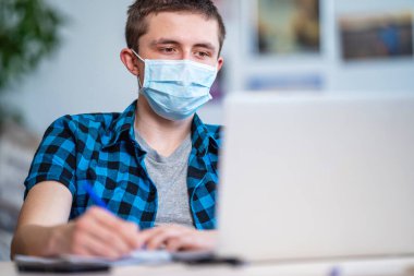 Koronavirüs karantinası sırasında laptopta ödevi olan maskeli ve kulaklıklı aktif bir genç. Sosyal uzaklık, kendini izole etme. Virüse bağlı uzaklık öğrenimi, COVID-19