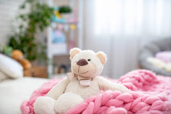 Cute white soft Teddy bear, sitting in a soft Merino blanket, large knitting, on the bed in the children\'s room. Childhood.