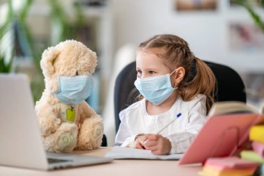 Maskeli küçük bir kız, oyuncak ayıyla ödev yapıyor, deftere yazıyor, dizüstü bilgisayar kullanıyor, e-öğrenme kavramları kullanıyor, karantina ve kendini tecrit sırasında. Covid-19 koronavirüs salgınından dolayı e-öğrenim.