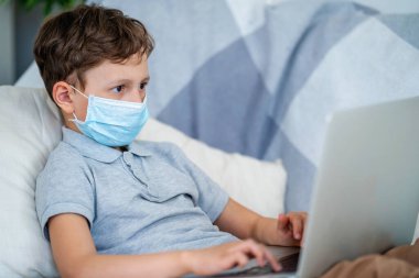 Koruyucu maskeli bir öğrenci elinde dizüstü bilgisayarla kanepede yatıyor. Çevrimiçi arkadaşlarla iletişim kuruyor. Antrenman videoları izliyor. Koronavirüs karantina döneminde uzaktan öğrenme.
