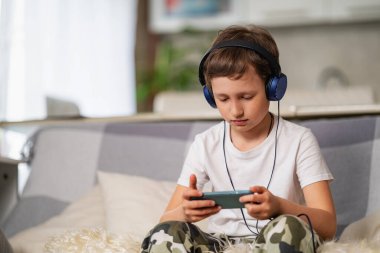 Kulaklıklı ve elinde akıllı telefon olan bir çocuk oturuyor. Cihazlara ve internete bağımlılık. Çocuk çevrimiçi oyunlar oynuyor ve salgın koronavirüs sırasında izolasyon döneminde boş zaman geçiriyor.