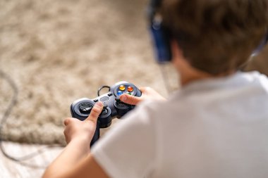 Kulaklıklı, elinde joystick olan, arkadan manzaralı bir çocuğun yakın çekimi. Bir çocuk video oyunları oynuyor. Boş zamanlarını, koronavirüs salgını sırasında kendini izole ederek geçirir..