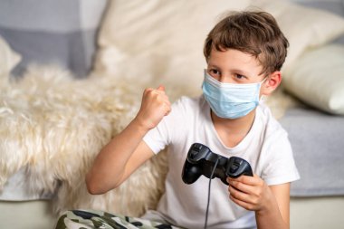 Maskeli neşeli çocuk, elinde joystickle bilgisayar oyunu kazanmaktan mutlu. Çocuk yalnızlık sırasında eğleniyor, koronavirüs salgını. Bilgisayar oyunlarına bağımlılık kavramı. Seçmeli olarak ele odaklan.