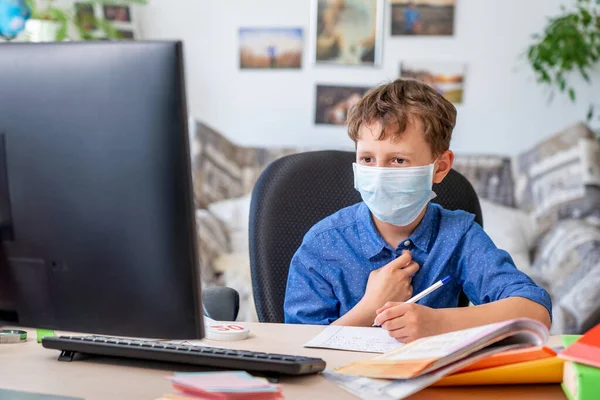 Boy in face mask with computer does his homework during a coronavirus quarantine. concept of online education. Social distance, self- isolation. Distance learning due to virus, flu, epidemic COVID-19