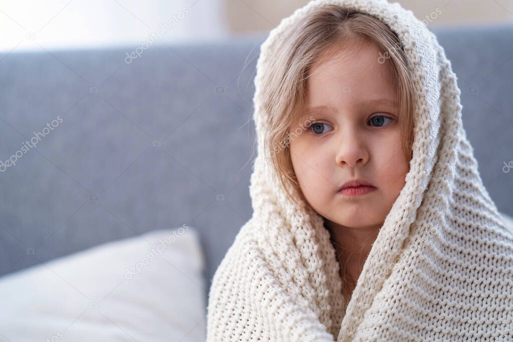 Portrait small blonde girl wrapped in blanket, sitting in bedroom after waking up in morning. child looks away and waits for his mother. baby in woolen blanket, it is at home, before going to bed.