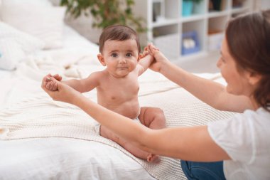 Yatakta destekli şirin bir bebek oturuyor. Anne, çocuk bakıcılığını destekliyor, ellerini yana doğru açıyor. Çocuk kendi kendine oturmaya başladı. Yeni beceri, destek ve bakım geliştirme kavramı.