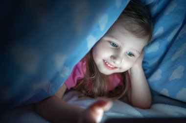 Sevimli küçük kız herkes uyurken akıllı telefonuyla battaniyenin altına saklanıyor. Çocuk gülümsüyor ve çizgi film izlerken eğleniyor. Çocukların yalnızlığı, internete ve cihazlara olan bağımlılığı.