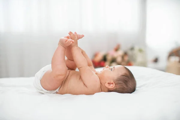 かわいい4ヶ月の赤ちゃんはベッドの上で彼の背中に横たわっていて 彼の手で遊んでいます 薄手のリネンでベッド 部屋は居心地が良く 光と花瓶の花がたくさんあります — ストック写真