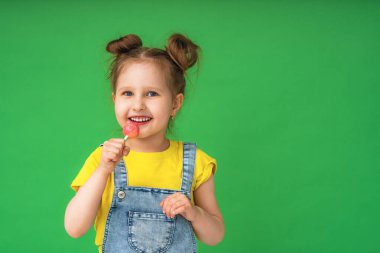 4 yaşında bir çocuk elinde lolipop tutuyor. Kameraya gülümsüyor, stüdyonun yeşil arka planında poz veriyor. Moda, güzellik, tatlılar, olumlu duygular, çocukların mutluluğu.