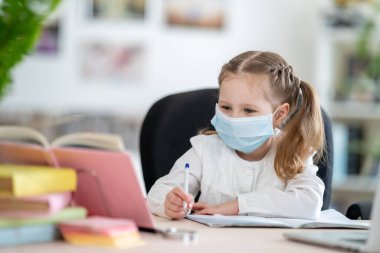 Maskeli tatlı bir kız, karantina ve kendini izole etme süresince dizüstü bilgisayar, e-öğrenme kavramları kullanarak ödev yapıyor. Covid-19 koronavirüs salgınıyla ilgili öğrenme.