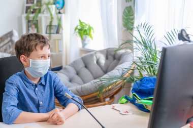 Yüzünde koruyucu maske olan bir çocuk, bilgisayarın başında oturuyor, karantina sırasında ödev yapıyor. Online eğitim kavramı. Sosyal uzaklık, kendini izole etme. Mesafe öğrenme, salgın hastalık