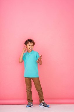 Dalgalı saçlı, Polo tişörtlü gülümseyen bir çocuk elinde bir akıllı telefon tutuyor ve pembe bir arka plana doğru işaret ediyor. Dijital teknolojiler, internet. Boşluğu kopyala