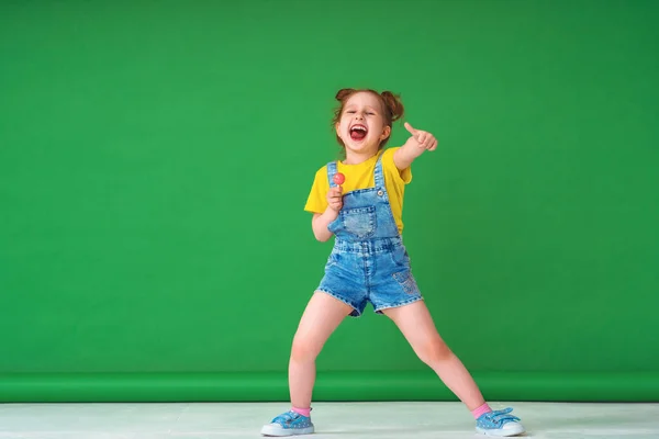 有趣的孩子表现出赞同的姿态 一个带棒棒糖的4岁女孩伸出了舌头 在工作室里一个绿色背景上顽皮的小女孩高兴地摆姿势 复制空间 — 图库照片