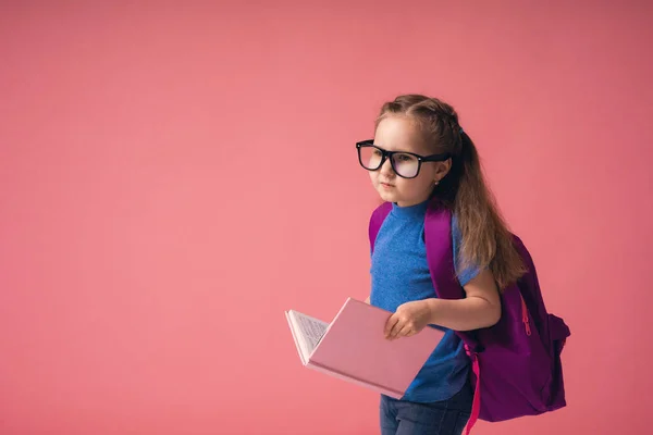 兴奋的小女孩 戴着眼镜 背着书包 拿着一本书去学校 空旷的空间被粉红的背景隔开 学校的教育理念 — 图库照片