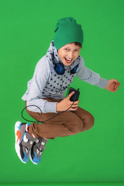 一个穿着牛仔裤 头罩和帽子的小男孩在绿色的背景上跳了起来 我看着摄像机 保持你的智能手机与连接耳机围住你的脖子 — 图库照片