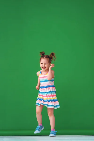 一个穿着五颜六色的条纹连衣裙跑着笑着的有趣滑稽的4岁女孩 工作室拍摄的绿色背景 幸福和无忧无虑的童年的概念 纵向方向 — 图库照片