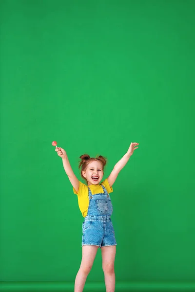 滑稽的孩子 高举双手在空中 一个带着棒棒糖的4岁女孩很有趣 感情上很快乐 在工作室里一个绿色背景上顽皮的小女孩高兴地摆姿势 — 图库照片