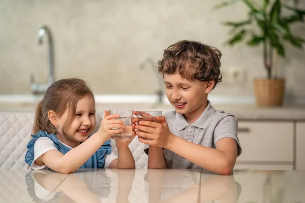Funny little children drink water in the kitchen at home. The boy and girl are smiling happily, holding a glass of clean water in their hands and looking at them. Water balance. Prevention of dehydration