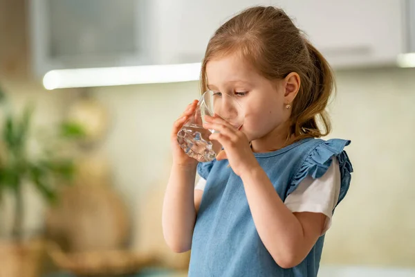Evdeki mutfakta su içen tatlı küçük kız. Çocuk bardağı temiz, filtrelenmiş içme suyunu elinde tutuyor ve susuzluğunu gidermek için zevkle içiyor. Su dengesi. Önleme kabızlığı