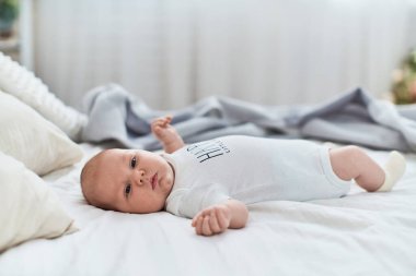 Beyaz tulumlu, çoraplı, sırt üstü yatıp başka tarafa bakan 4 aylık tatlı bir bebek. Hafif çarşaflı, battaniyeli ve yastıklı bir yatak. Oda rahat ve aydınlık..
