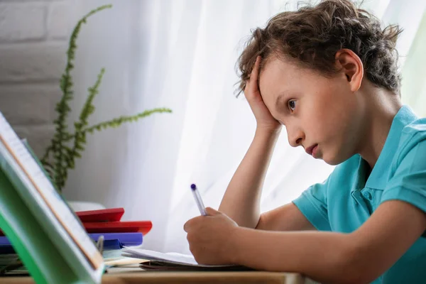 悲しい少年は彼の頭の下に手を置き 本を見て 机に座って宿題を読みます 学校に戻る 子供は勉強に疲れている 学年末 子供は課題を理解していません — ストック写真