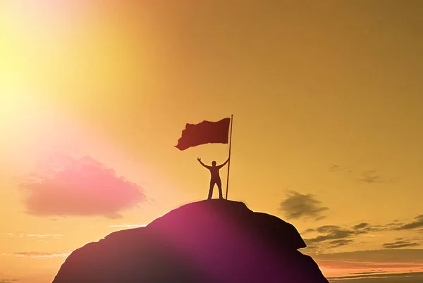 Alta realização, silhuetas de homens, bandeira da vitória no topo da montanha, mãos para cima. Um homem no topo de uma montanha. Design conceitual. Contra o céu dramático com nuvens ao pôr do sol . — Fotografia de Stock