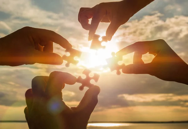 Vier Hände versuchen, ein Puzzleteil mit dem Hintergrund eines Sonnenuntergangs zu verbinden. ein Puzzle in der Hand gegen das Sonnenlicht. Ein Teil des Ganzen. Stockbild