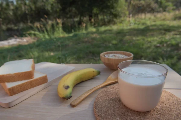 パンと木製のテーブルの朝食のバナナとミルクのガラス。朝の日差し. — ストック写真