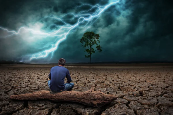 Homem sentado em madeira em terra para o chão seco rachado e árvore grande. Com uma tempestade relâmpago — Fotografia de Stock