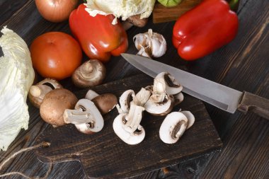 Karanlık bir tahta üzerinde kesilmiş taze mantarlar, bir bıçak, olgun kırmızı domates, dolma biber, ahşap bir arka planda marul yaprakları, sebzeler.