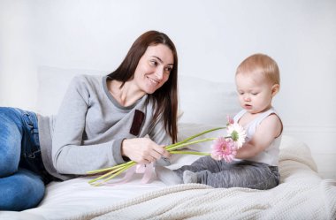 Anne ve kızı pembe çiçeklerle beyaz bir yatakta oturuyor, çocuklu bir kadın.