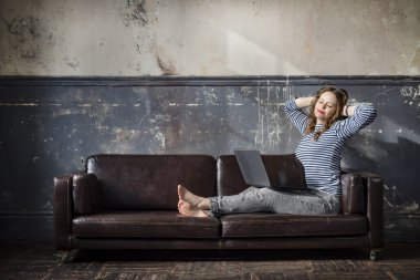 Bir beyaz kadın kot pantolon ve yelek giymiş eski bir duvarın arkasında oturuyor dizüstü bilgisayarlı ve yüzünde bir gülümsemeyle, güneş ışınları