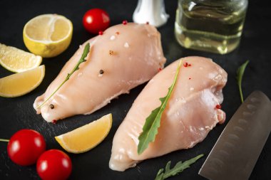 Mutfak tahtasında çiğ tavuk filetosu, domates, limon, otlar, siyah arka planda et yakın plan.