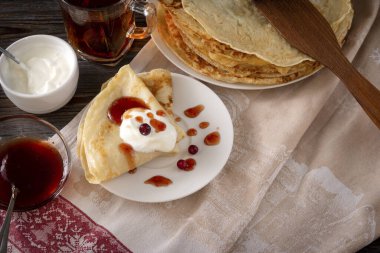 Böğürtlenli, kızılcıklı, ekşi kremalı ve reçelli bir sürü krep. Koyu ahşap arka planda bir havlunun üzerinde bir kupa siyah çay.