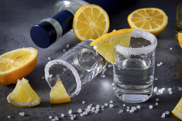 2 рюмки серебряной текилы с солью и ломтиками лимона на сером фоне, бутылки на заднем плане, крепкий алкогольный напиток, текила, водка
,